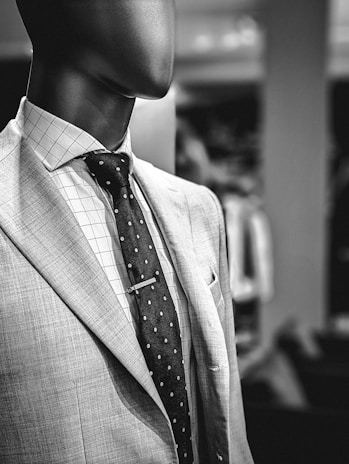 A mannequin is dressed in a stylish suit, featuring a light-colored jacket with a subtle texture, a white dress shirt with a grid pattern, and a dark tie adorned with small polka dots. The focus is on the upper body and the outfit's classic elements.