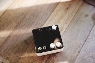 A black electronic device with several metallic knobs and connectors is placed on a wooden surface. The sunlight casts gentle shadows and highlights on the wooden texture, creating a warm and mellow atmosphere.