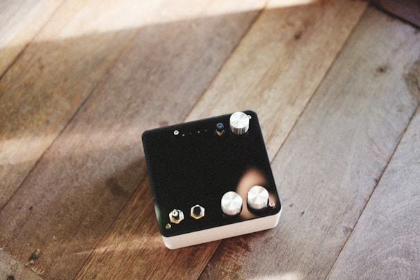 A black electronic device with several metallic knobs and connectors is placed on a wooden surface. The sunlight casts gentle shadows and highlights on the wooden texture, creating a warm and mellow atmosphere.