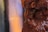 Slices of raw meat stacked closely together with a glossy texture, showing a rich maroon color. The background is a blurred blend of purple and orange shades.