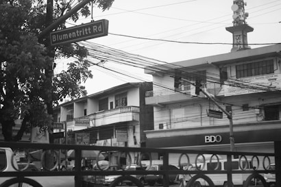 A street scene with a road sign labeled 'Blumentritt Rd' next to a tree. The area is surrounded by buildings, including a security training center and a BDO branch. Cars and pedestrians populate the street, with overhead electrical wires visibly crisscrossing the scene. The atmosphere is urban and slightly overcast.