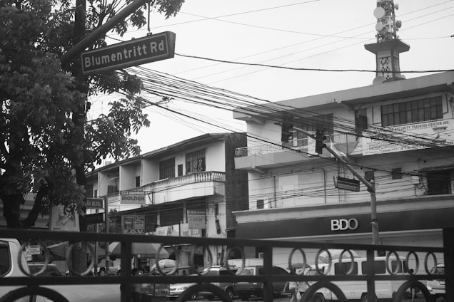 A street scene with a road sign labeled 'Blumentritt Rd' next to a tree. The area is surrounded by buildings, including a security training center and a BDO branch. Cars and pedestrians populate the street, with overhead electrical wires visibly crisscrossing the scene. The atmosphere is urban and slightly overcast.