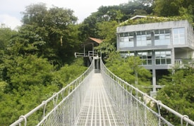 A metal suspension bridge extends over lush green foliage towards a modern glass-walled building nestled among dense trees. The structure is elevated, with a pathway allowing passage over the vibrant greenery.