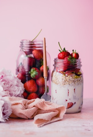Two glass jars are displayed on a light pink background. The first jar is filled with fresh strawberries, while the second contains a layered parfait with strawberries, yogurt, and granola. A pink napkin and a spoon are placed nearby, alongside peony flowers.