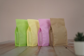Four stand-up pouches in pastel colors including light green, yellow, pink, and beige are lined up on a wooden surface against a soft beige background. A small plant in a white pot is partially visible in the bottom right corner.
