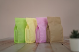 Four stand-up pouches in pastel colors including light green, yellow, pink, and beige are lined up on a wooden surface against a soft beige background. A small plant in a white pot is partially visible in the bottom right corner.