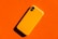 A bright yellow smartphone case lies on a vibrant orange background, casting a shadow due to angled lighting. The phone case is minimalist in design and features rounded edges.
