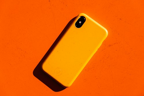 A bright yellow smartphone case lies on a vibrant orange background, casting a shadow due to angled lighting. The phone case is minimalist in design and features rounded edges.