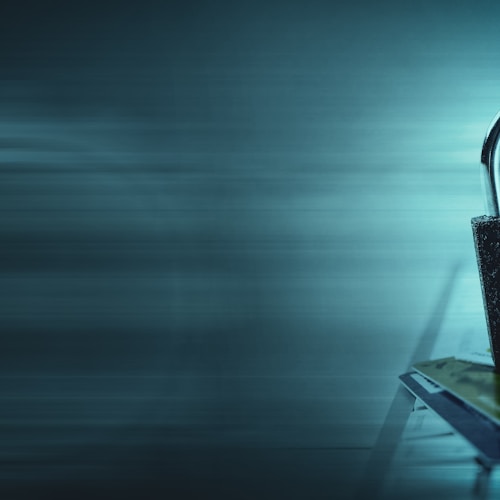 A padlock sits atop a stack of credit cards on a keyboard, with a bluish hue and digital streaks in the background, suggesting themes of digital security and privacy.