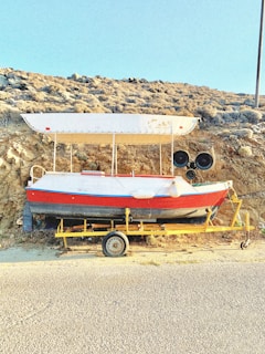A small boat is situated on a yellow trailer on the side of a road, with a rocky and dry hillside in the background. The boat has a white canopy and is painted red and white with a blue stripe. There are two circular, black speakers attached near the front.