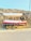 A small boat is situated on a yellow trailer on the side of a road, with a rocky and dry hillside in the background. The boat has a white canopy and is painted red and white with a blue stripe. There are two circular, black speakers attached near the front.