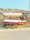 A small boat is situated on a yellow trailer on the side of a road, with a rocky and dry hillside in the background. The boat has a white canopy and is painted red and white with a blue stripe. There are two circular, black speakers attached near the front.