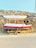 A small boat is situated on a yellow trailer on the side of a road, with a rocky and dry hillside in the background. The boat has a white canopy and is painted red and white with a blue stripe. There are two circular, black speakers attached near the front.