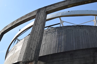 A close-up view of a large, circular concrete structure with reinforced beams and metal support elements. The architecture showcases an industrial design with exposed concrete and metal contrasting against a clear blue sky in the background.