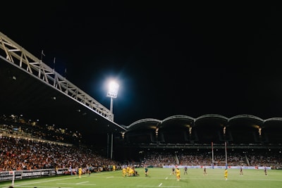 A nighttime rugby match with players on the field and spectators filling the stadium seats under bright floodlights. The stadium has a modern design with an intricate roof structure and illumination highlighting the energetic atmosphere of the event.