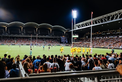 A large stadium filled with spectators watching a nighttime rugby match. The players are wearing yellow jerseys and the illuminated scoreboard is visible in the background. The stadium has a modern design with overhead lights.