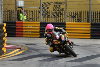A motorcyclist wearing a bright pink helmet is racing on a track. The motorcycle leans sharply as it navigates a curve, displaying the number 3 prominently on the front. The track is surrounded by yellow and black barriers, with a marked exit visible in the background. A person in a neon vest stands behind the barriers, observing the race. The scene is set against a backdrop featuring the words 'Grand Prix Macau'.