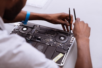 Hands of a person working on the internal components of a laptop, including the fan and circuit board, using two precision tools. The person is wearing an anti-static wrist strap.