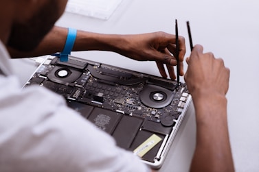 Hands of a person working on the internal components of a laptop, including the fan and circuit board, using two precision tools. The person is wearing an anti-static wrist strap.