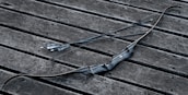 A black archery bow and two arrows are placed on a weathered wooden surface. The bow has a sleek, modern design with textured grip, and the arrows are straight with pointed tips and feathered ends.
