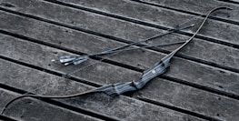 A black archery bow and two arrows are placed on a weathered wooden surface. The bow has a sleek, modern design with textured grip, and the arrows are straight with pointed tips and feathered ends.