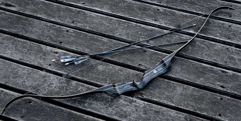 A black archery bow and two arrows are placed on a weathered wooden surface. The bow has a sleek, modern design with textured grip, and the arrows are straight with pointed tips and feathered ends.