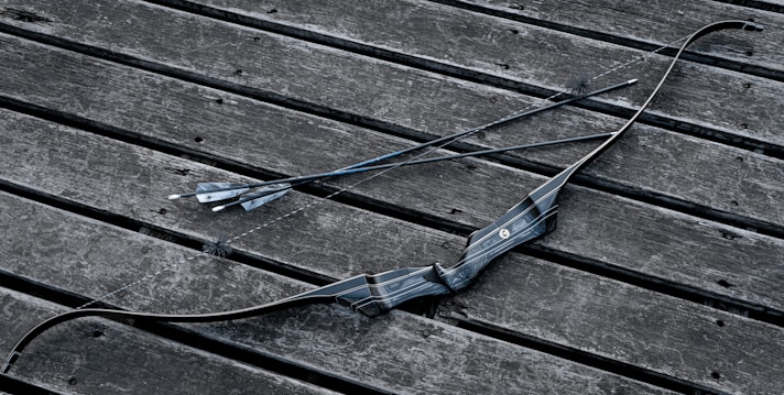 A black archery bow and two arrows are placed on a weathered wooden surface. The bow has a sleek, modern design with textured grip, and the arrows are straight with pointed tips and feathered ends.