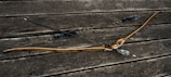 A wooden bow and two arrows with fletching rest on a weathered wooden surface. The bow has a natural wood finish with a dark grip area, while the arrows have dark shafts and are outfitted with fluffy dampeners.