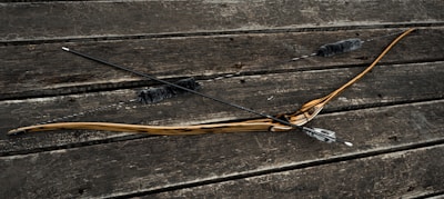 A wooden bow and two arrows with fletching rest on a weathered wooden surface. The bow has a natural wood finish with a dark grip area, while the arrows have dark shafts and are outfitted with fluffy dampeners.