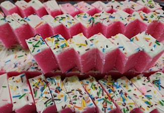 A collection of brightly colored pink and white rectangular sweets, topped with a layer of white icing and multi-colored sprinkles. The sweets are neatly arranged in stacked rows.