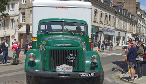 A vintage green Volvo truck is driving down a street lined with old stone buildings. People are gathered on the sidewalk, some taking photos, as the truck passes by. The street has a charming, historical town vibe with people enjoying the event.