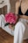 An individual with a tattoo on their arm is seated on the floor, holding a vibrant pink peony flower with green leaves. They are wearing a black top and white, slightly torn jeans. A glimpse of wooden flooring and a white piece of furniture is visible in the background.