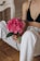 An individual with a tattoo on their arm is seated on the floor, holding a vibrant pink peony flower with green leaves. They are wearing a black top and white, slightly torn jeans. A glimpse of wooden flooring and a white piece of furniture is visible in the background.