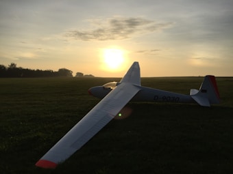 A glider rests on a grassy field during sunset, casting a long shadow. The sky is filled with warm hues of orange and yellow, while sparse clouds create a serene atmosphere. The glider's structure is sleek and white, with a distinctive red tip.