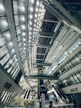 A spacious, modern interior with high ceilings and a futuristic architectural design. The interior is illuminated by rows of bright lights, creating a symmetrical pattern across the ceiling. Multiple levels with glass railings and elevators are visible, along with stairs and escalators. The structure is primarily composed of glass, steel, and concrete.
