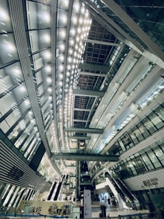 A spacious, modern interior with high ceilings and a futuristic architectural design. The interior is illuminated by rows of bright lights, creating a symmetrical pattern across the ceiling. Multiple levels with glass railings and elevators are visible, along with stairs and escalators. The structure is primarily composed of glass, steel, and concrete.