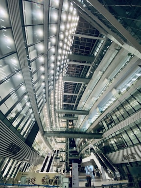 A spacious, modern interior with high ceilings and a futuristic architectural design. The interior is illuminated by rows of bright lights, creating a symmetrical pattern across the ceiling. Multiple levels with glass railings and elevators are visible, along with stairs and escalators. The structure is primarily composed of glass, steel, and concrete.
