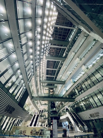 A spacious, modern interior with high ceilings and a futuristic architectural design. The interior is illuminated by rows of bright lights, creating a symmetrical pattern across the ceiling. Multiple levels with glass railings and elevators are visible, along with stairs and escalators. The structure is primarily composed of glass, steel, and concrete.
