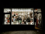 A store display filled with various Halloween costumes and decorations, featuring multiple mannequin heads wearing different masks and wigs. The window is brightly lit, showcasing colorful costumes, spooky masks, and accessories with a slightly eerie ambiance.