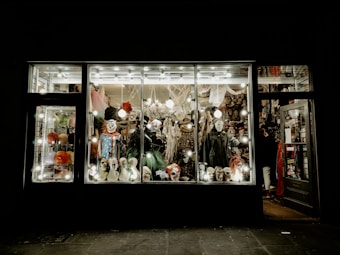 A store display filled with various Halloween costumes and decorations, featuring multiple mannequin heads wearing different masks and wigs. The window is brightly lit, showcasing colorful costumes, spooky masks, and accessories with a slightly eerie ambiance.
