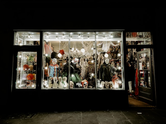 A store display filled with various Halloween costumes and decorations, featuring multiple mannequin heads wearing different masks and wigs. The window is brightly lit, showcasing colorful costumes, spooky masks, and accessories with a slightly eerie ambiance.