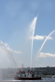 A fireboat is spraying large arcs of water into the air. The vessel, equipped with multiple water cannons, is positioned on a body of water with a clear sky above. In the background, there are industrial buildings lining the waterfront.