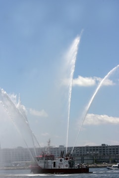 A fireboat is spraying large arcs of water into the air. The vessel, equipped with multiple water cannons, is positioned on a body of water with a clear sky above. In the background, there are industrial buildings lining the waterfront.