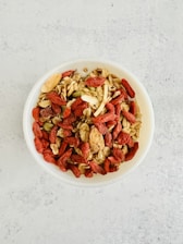 A bowl filled with a mixture of various nuts, seeds, and dried fruits, including almonds, walnuts, pumpkin seeds, and red goji berries. The bowl is placed on a light-colored, textured surface.