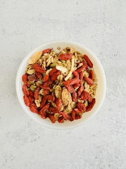 A bowl filled with a mixture of various nuts, seeds, and dried fruits, including almonds, walnuts, pumpkin seeds, and red goji berries. The bowl is placed on a light-colored, textured surface.