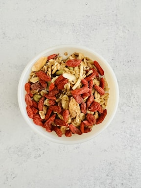 A bowl filled with a mixture of various nuts, seeds, and dried fruits, including almonds, walnuts, pumpkin seeds, and red goji berries. The bowl is placed on a light-colored, textured surface.
