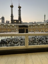 A large crowd of people in white garments gathered around the Kaaba, the central black structure, in the courtyard of the Grand Mosque. Tall cranes and under-construction buildings are visible in the background, along with illuminated minarets and domes.