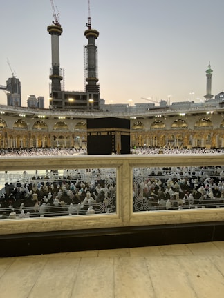 A large crowd of people in white garments gathered around the Kaaba, the central black structure, in the courtyard of the Grand Mosque. Tall cranes and under-construction buildings are visible in the background, along with illuminated minarets and domes.