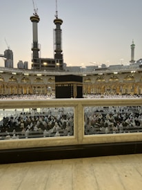 A large crowd of people in white garments gathered around the Kaaba, the central black structure, in the courtyard of the Grand Mosque. Tall cranes and under-construction buildings are visible in the background, along with illuminated minarets and domes.