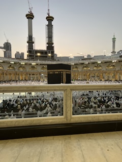 A large crowd of people in white garments gathered around the Kaaba, the central black structure, in the courtyard of the Grand Mosque. Tall cranes and under-construction buildings are visible in the background, along with illuminated minarets and domes.